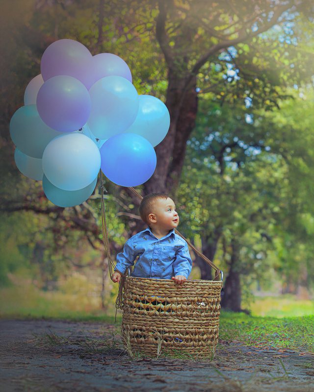 Baby in a basket outdoors beneath pastel balloons.