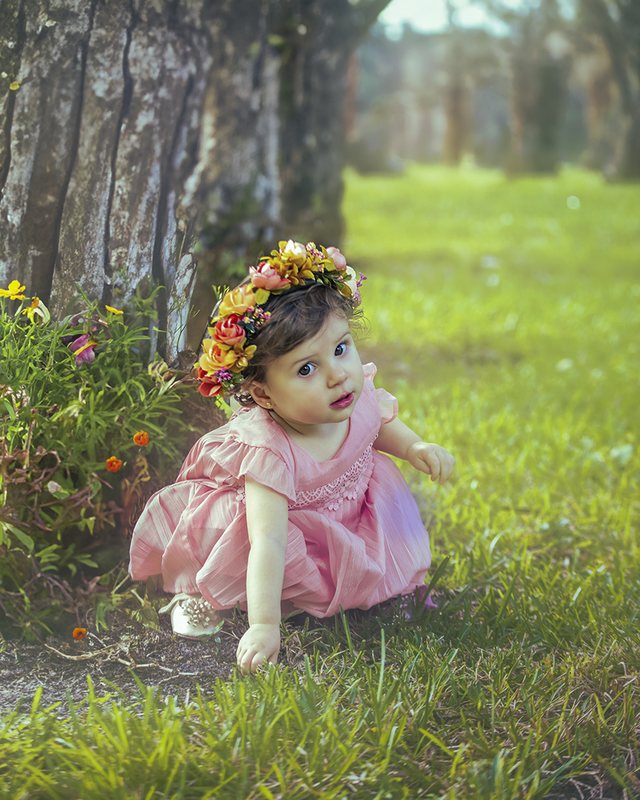 Baby wearing pink and a flower crown beside a tree.