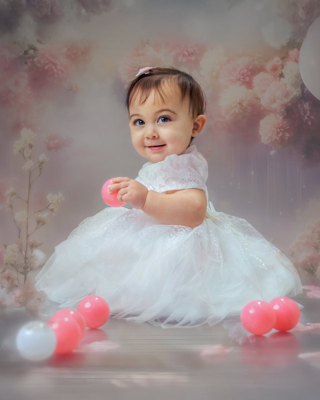 Baby in a white dress seated with pink and white decorations.