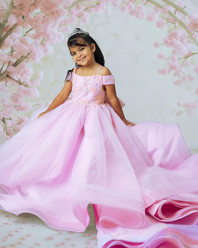 Girl in a pink gown and tiara seated against a floral backdrop.