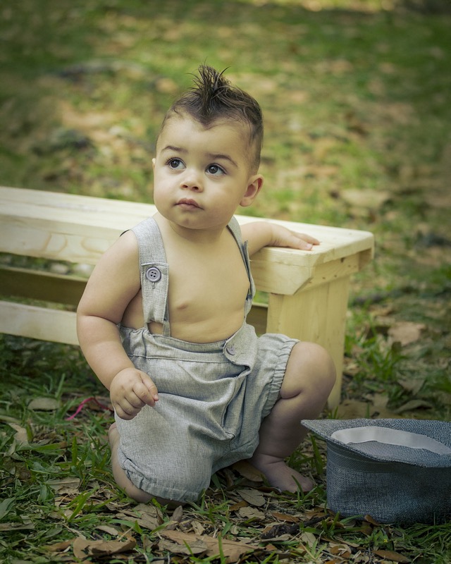 Baby in gray overalls kneeling beside a wooden tub outdoors.