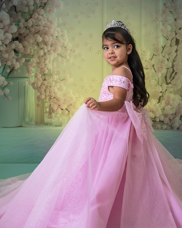 Girl in a pink gown and tiara posing beside a floral backdrop.