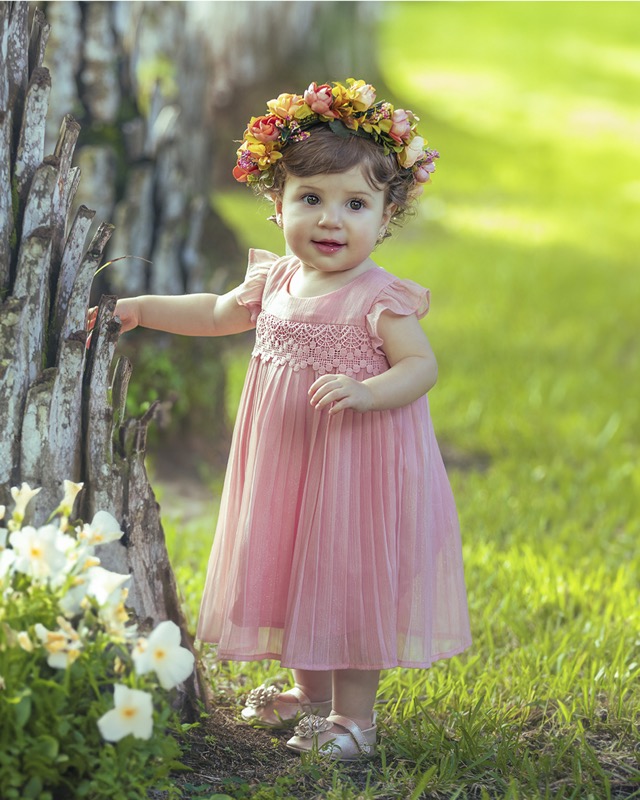 Young child in a pink dress and colorful flower crown beside a tree.