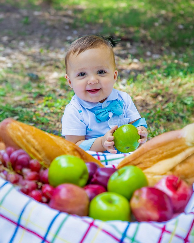 Smiling baby wearing a blue bow tie seated on a picnic blanket with fruit and bread.