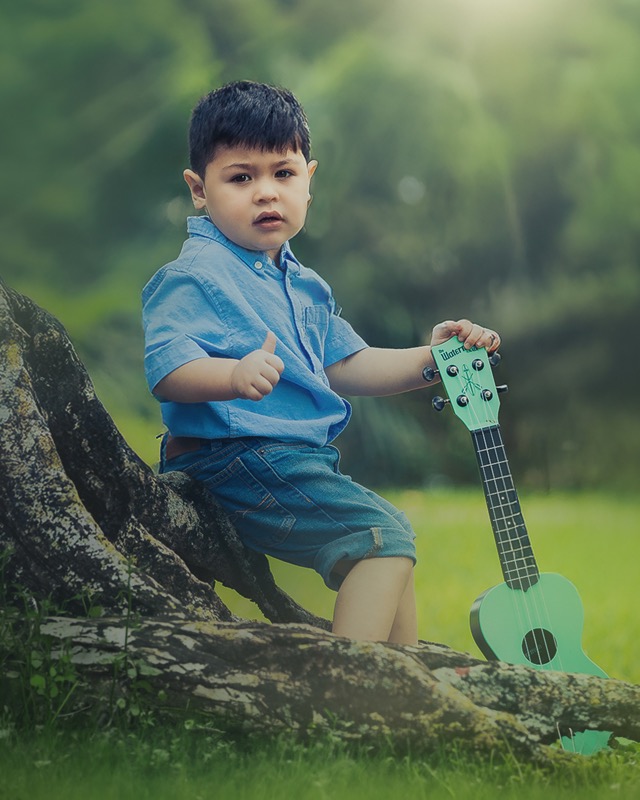 Young child in denim seated on a tree branch beside a green ukulele.