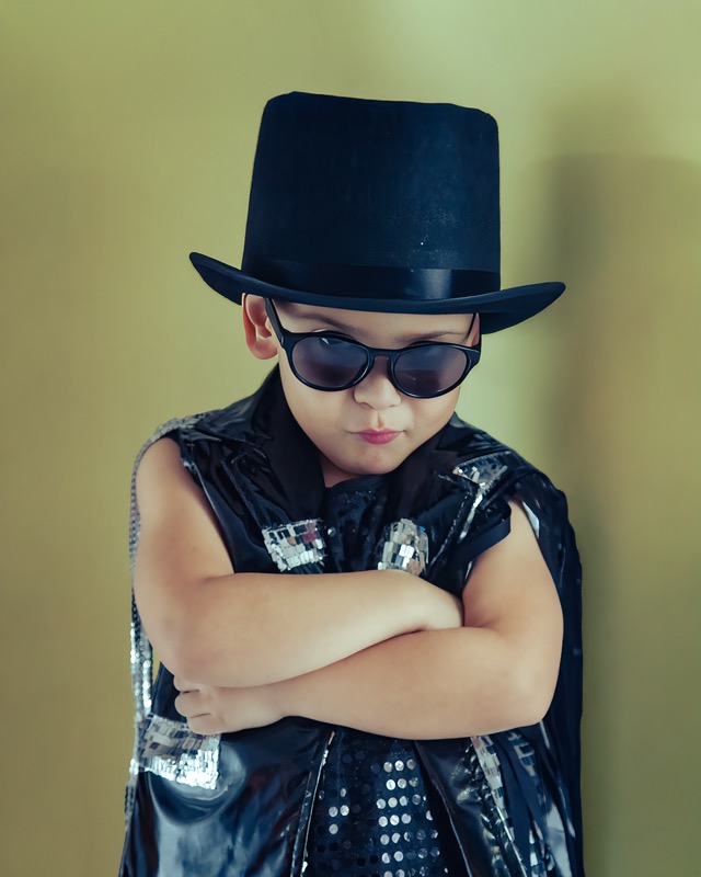 Young child wearing a black top hat, sunglasses and a shiny black vest.