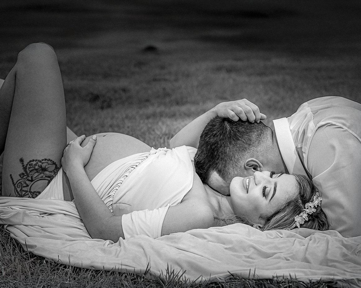 Black-and-white maternity portrait of an expectant mother smiling as her partner kisses her neck.