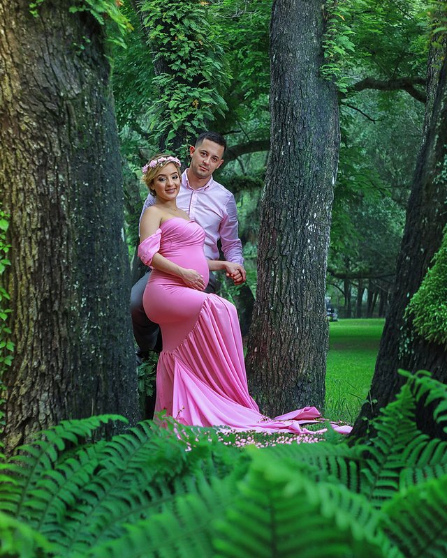 Couple posing among trees, with the expectant mother wearing a pink dress.