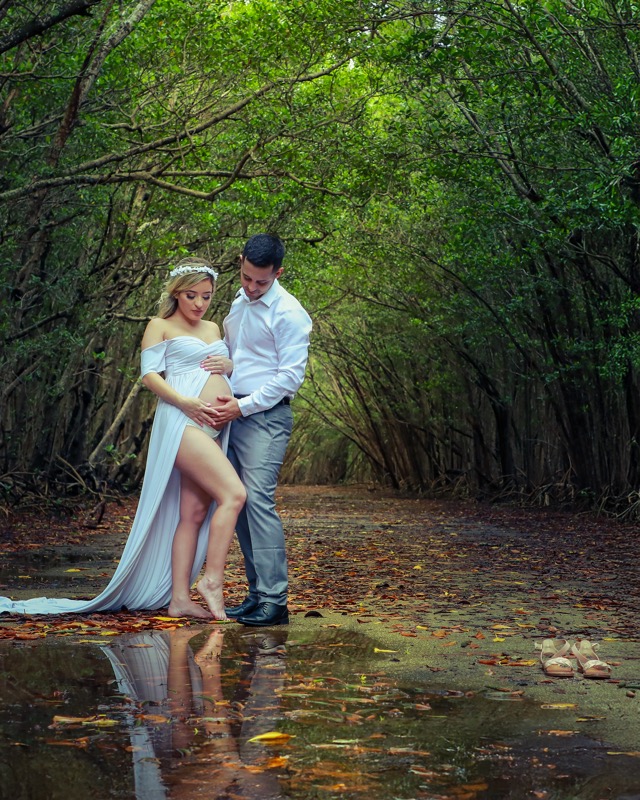 Expectant couple beneath a canopy of trees beside a puddle reflecting their image.