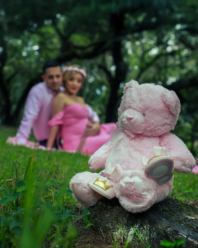 Pink teddy bear and tiny shoes on the grass, with an expectant couple in the background.