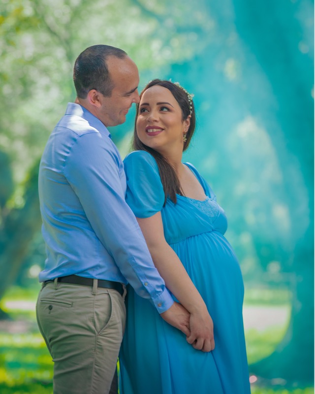 Expectant mother in a blue dress embraced by her partner beside a tree.